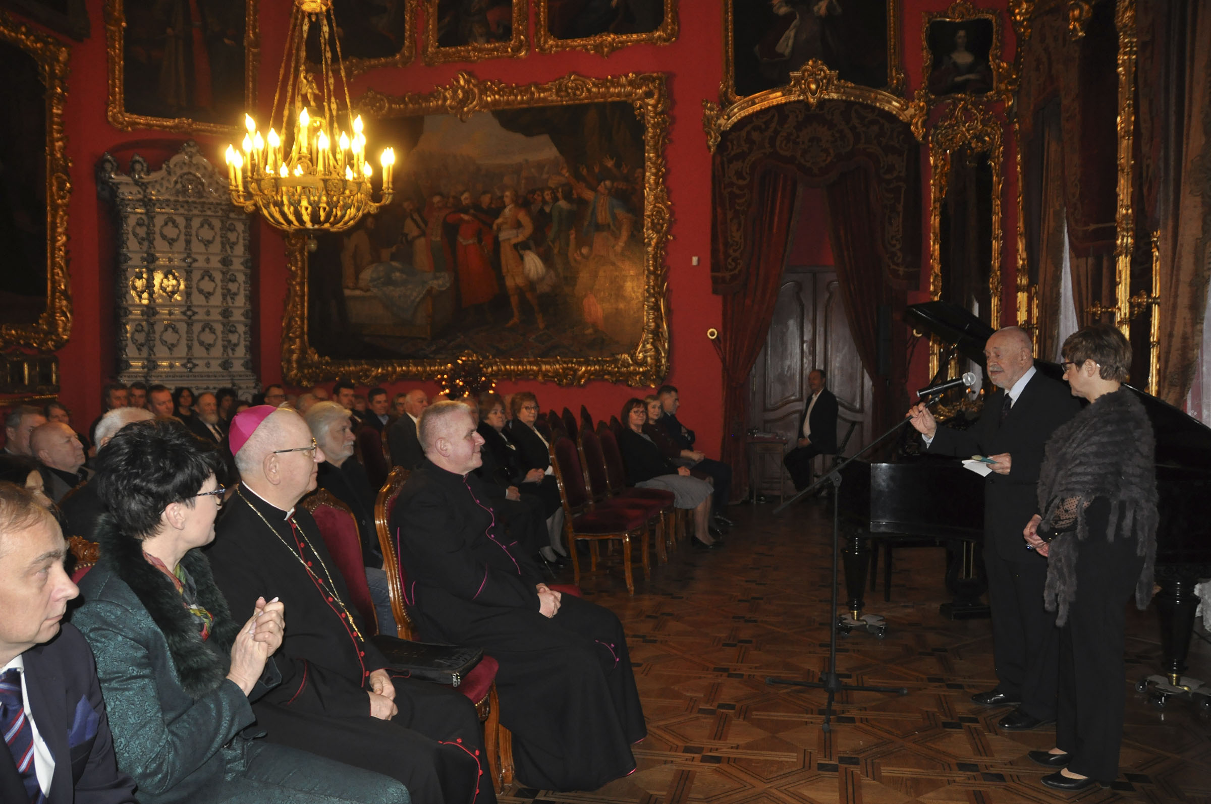 Spotkanie noworoczne Towarzystwa Przyjaciół Muzeum Fotografia. Duże pomieszczenie pomalowane na czerwono, nastrojowo oświetlone. Na ścianach w bogato rzeźbionych i złoconych ramach wiszą w równych odległościach obrazy. W głębi okazały piec kaflowy. Po lewej stronie na obitych bordowym aksamitem krzesłach siedzą zasłuchani ludzie. Po prawej stronie mówiący do mikrofonu starszy mężczyzna, przy nim kobieta w okularach z narzuconą ciemnoszarą etolą z miękkiego futra.
