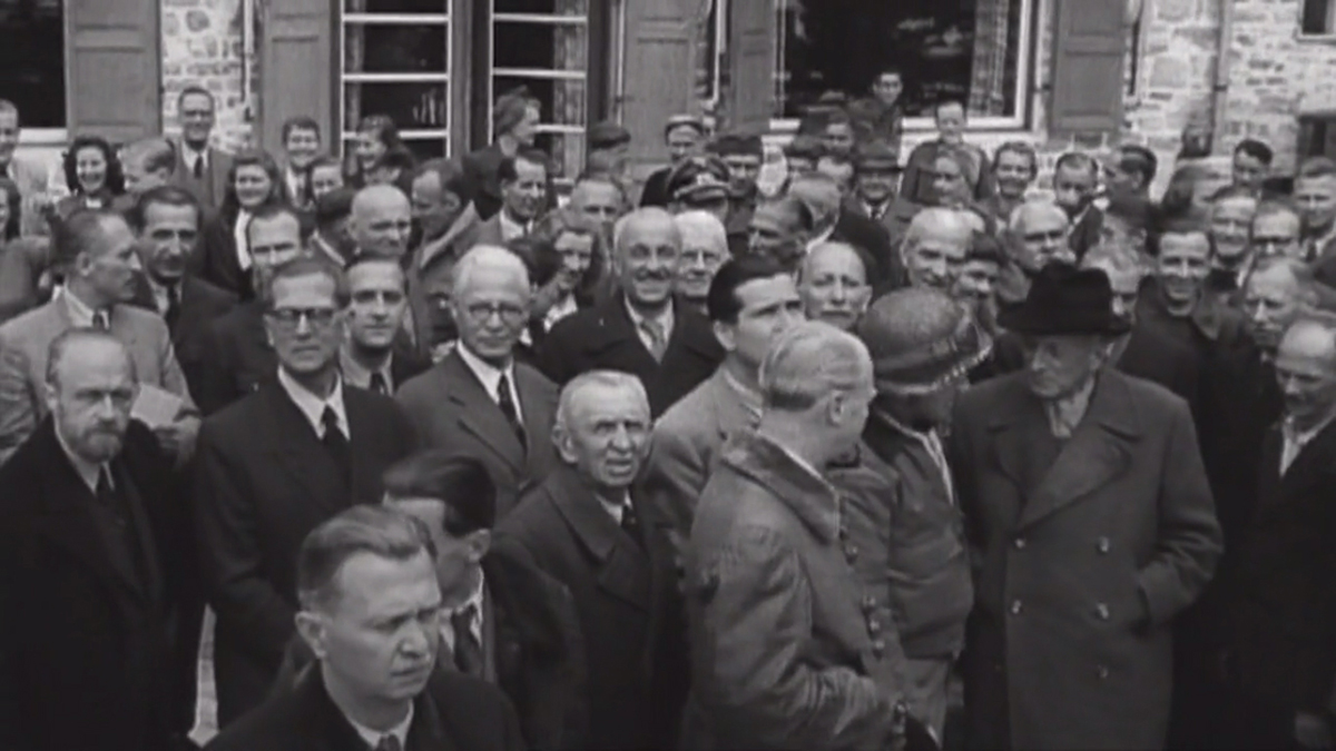 Kadr z dokumentu fabularyzowanego „Wir Geiseln der SS”,2015, https://vimeo.com/groups/269270/videos/136490059 Aleksander Zamoyski, po uwolnieniu, z lewej, w jasnej marynarce. Kadr z dokumentu fabularyzowanego „Wir Geiseln der SS”, (Jesteśmy zakładnikami SS). Duża grupa ludzi na tle budynku z kamienną fasadą i okiennicami. Postacie z bliższego planu skierowane twarzą w stronę fotografa. Po prawej stronie u dołu trzech mężczyzn zajętych rozmową.