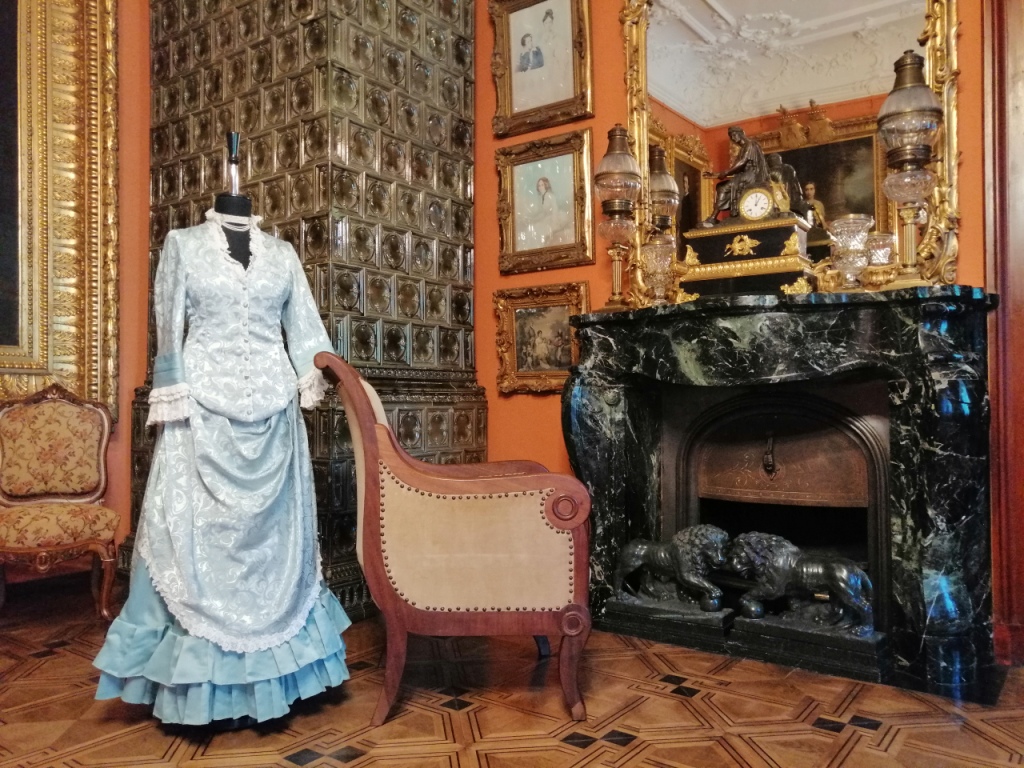 Wystawa "Modna w każdym wieku" Fotografia. Fragment pomieszczenia. Przy jasnym fotelu ustawionym przed kominkiem z zielonego marmuru dziewiętnastowieczna stylizowana suknia zaprezentowana na manekinie. W tle wysoki piec z oliwkowych kafli. Suknia składa się ze stanika i dwuczęściowej spódnicy. Spodnia jej część uszyta jest z jednobarwnego seledynowego materiału wykończonego u dołu dwurzędową, plisowaną falbanką. Górna warstwa, drapowana na biodrach wykonana jest z jaśniejszego materiału z kwiatową aplikacją. Z tej samej tkaniny uszyty jest też stanik. Długie rękawy zakończone są delikatną koronką. Tak samo obszyty jest dekolt i górna warstwa spódnicy.
