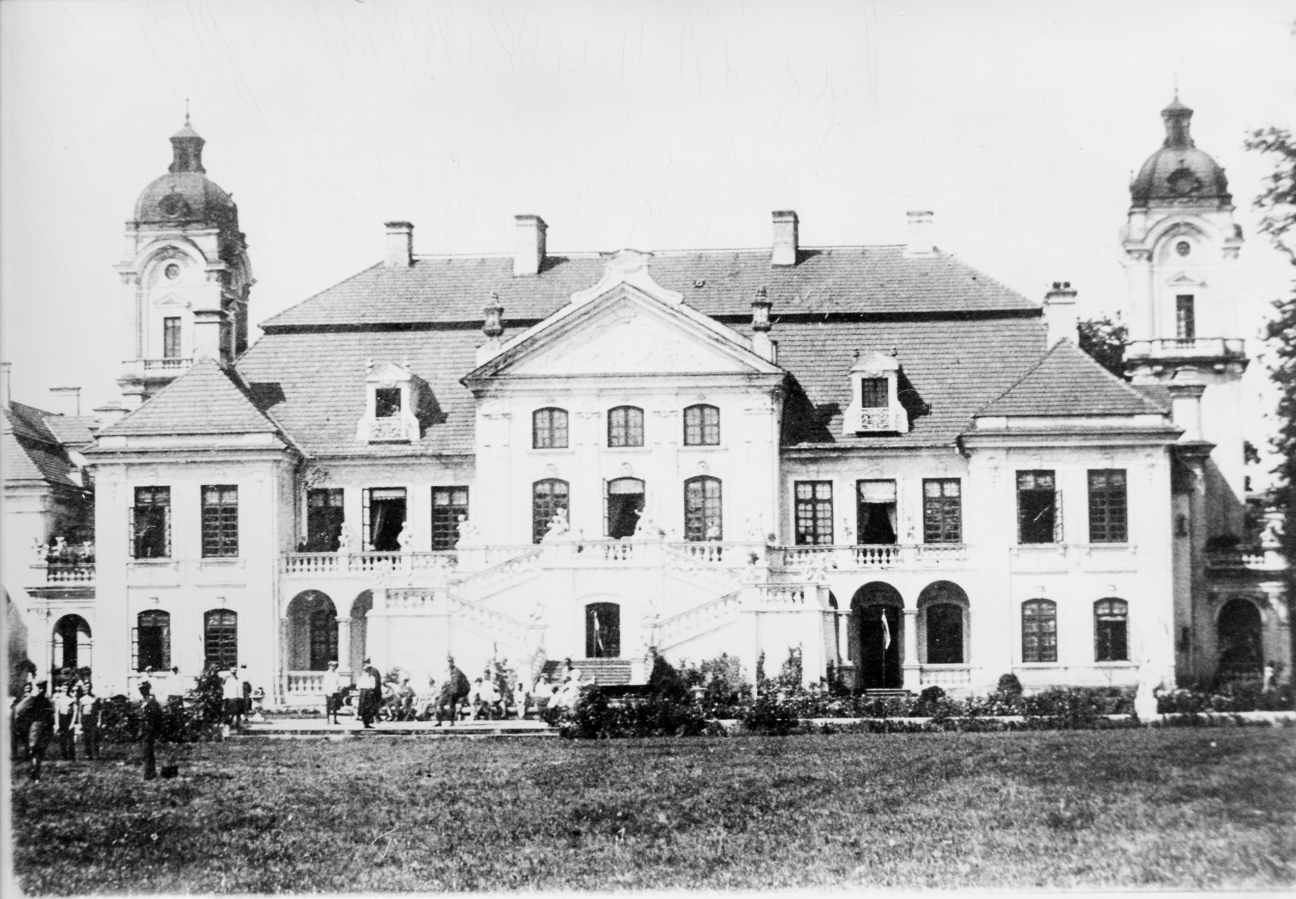 Pałac w Kozłówce - elewacja ogrodowa, okres międzywojenny Czarno-biała fotografia. Okazały jednopiętrowy pałac z trzema ryzalitami: środkowym i bocznymi. Do środkowego ryzalitu dostawiony olbrzymi taras z podcieniami i schodami do ogrodu. Dach łamany, mansardowy, pokryty dachówką. Przy krótszych bokach pałacu widoczne wieże z dachem w kształcie kopuły. Górują nad pałacem Przed pałacem w małych grupkach zgromadzeni ludzie.