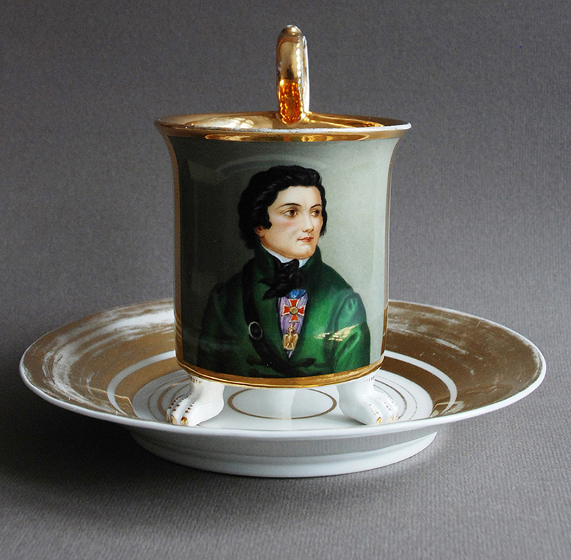 Filiżanka z wizerunkiem Tadeusza Kościuszki Fotografia. Filiżanka z miniaturą Tadeusza Kościuszki, ze spodkiem. Berlin, początek dwudziestego wieku. Porcelana, kalkomania, farby naszkliwne, złocenia. Wysokość 10 centymetrów.
Filiżanka na trzech nóżkach w formie lwich łap. Na szarozielonym tle Kościuszko w ujęciu do pasa. Naczelnik ubrany w zieloną, zdobioną pasmanteryjnym zapięciem, sukmanę i białą koszulę z czarną krawatką. Na piersi krzyż Virtuti Militari i amerykański order Cyncynata. Przez pierś przełożony pas ładownicy. Wnętrze filiżanki złocone. Spodek ze złoconym kołnierzem.
