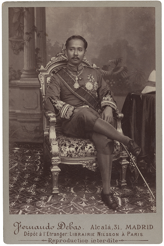 Czulalongkorn (Rama V), król Syjamu (Tajlandia), fot. Fernando Debas, Madryt, lata 80. XIX w. Dojrzały mężczyzna o azjatyckich rysach twarzy i smagłej karnacji, z ciemnymi włosami i wąsami, siedzący w fotelu z nogą założoną na nogę, w całej postaci na wprost. Ubrany jest w mundur z bogatymi aplikacjami na rękawach i stojącym kołnierzu, przepasany szarfą, z przypiętymi czterema gwiazdami orderowymi i zawieszonym na szyi orderem na łańcuchu. Na nogach ma ciemne pumpy, pończochy i sznurowane pantofle. Przy lewym boku przypięta szpada. Z prawej strony nakryty serwetą stół, na nim hełm korkowy. W tle ekran z dwiema kolumnami i pejzażem, na podłodze wzorzysty dywan. Fotografia w kolorze sepii.