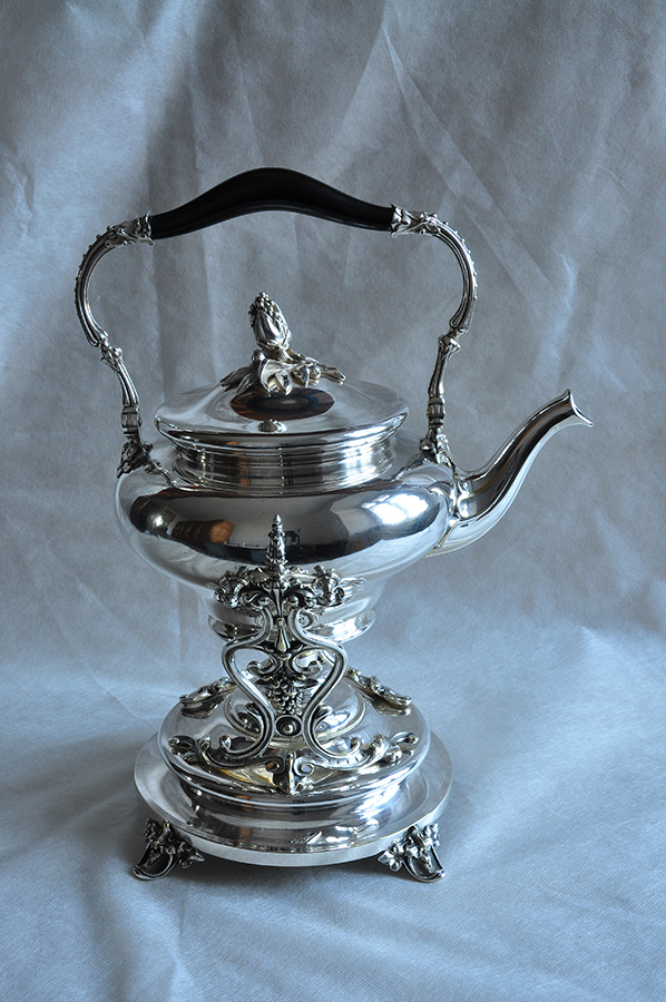 Buliera, Francja, Paryż, Christofle, 1865; metal, odlew, wytłaczanie, srebrzenie, heban; wys. 38,5 cm; zbiory Muzeum Zamoyskich w Kozłówce, nr inw. MPK/ZM/1400 Fotografia przedstawia czajnik na podstawie z podgrzewaczem. Obydwa elementy w kolorze srebrnym. Czajnik o pękatym korpusie, w kształcie gruszkowatym, z prawej strony esowato wygięty dzióbek. Kabłąkowaty uchwyt uformowany z dwóch części dekorowanych listkami połączonych środkowym drewnianym fragmentem w kolorze ciemnym. Pokrywka wysklepiona, zwieńczona stylizowanym ulistnionym pąkiem. Okrągła podstawa podgrzewacza, widoczne dwie nóżki w kształcie wywiniętych ulistnionych kartuszy. Palnik umieszczony pośrodku, zasłonięty ornamentem kandelabrowym z ułożonych symetrycznie, wygiętych rozgałęzionych łodyg przechodzących przy podstawie w roccaile ujęte liśćmi akantu. Powyżej w wić wkomponowane kwiaty, kiść winogron oraz motywy zgeometryzowane.