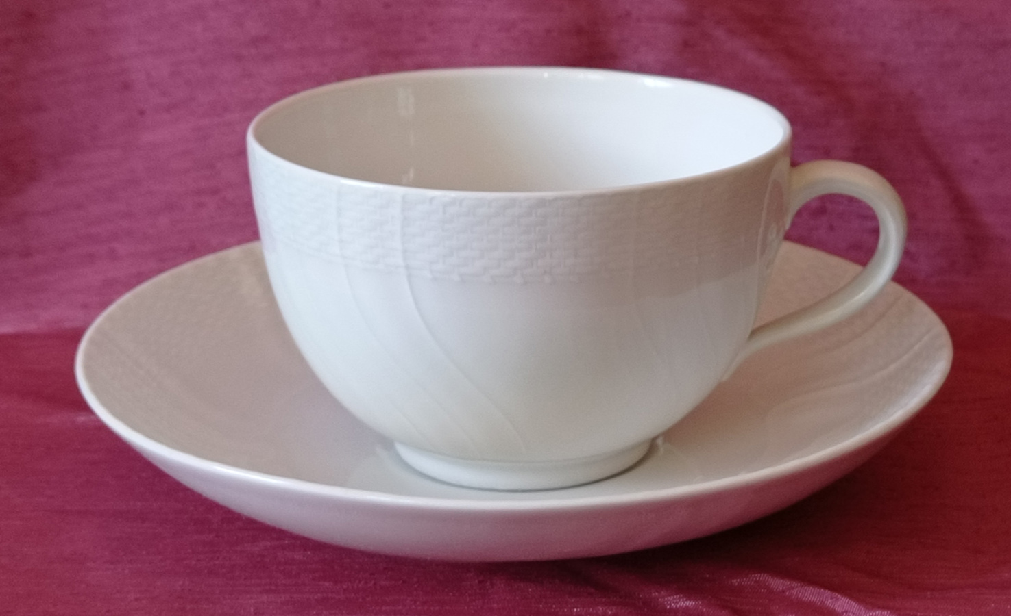 Filiżanka ze spodkiem (z serwisu stołowego), Niemcy, Berlin, Königliche Porzellan Manufaktur, 1913 -1918; porcelana; wys. 6,6 cm, średn. 10,4 cm (filiżanka), wys. 3,3 cm, średn. 17 cm (spodek); zbiory Muzeum Zamoyskich w Kozłówce, nr inw. MPK/CS/880 (filiżanka), MPK/CS/909 (spodek) Fotografia przedstawia zestaw: filiżanka ze spodkiem w kolorze białym na czerwonym tle. Filiżanka na niskiej taśmowej podstawie, półkulista, u góry pasek z dekoracją przypominającą plecionkę z wikliny, na całości równolegle ułożone, pionowe, esowate, wypukłe linie. Ten sam wzór powtórzony na spodku.