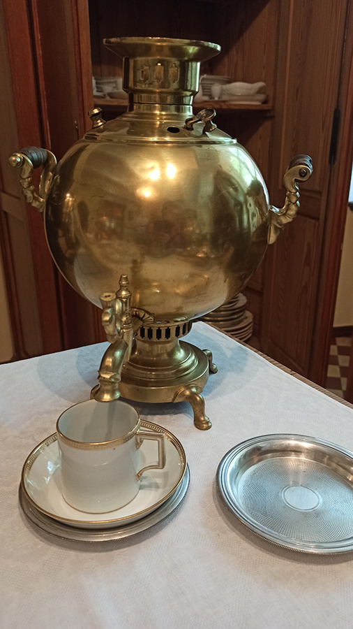 Samowar, Rosja, początek XX wieku; mosiądz, odlew; wys. 36 cm; zbiory Muzeum Zamoyskich w Kozłówce, nr inw. MPK/ZM/131 Fotografia przedstawia fragment stołu. Na nim na pierwszym planie, z lewej strony biała filiżanka z uszkiem w kolorze złotym i dekoracyjnym paskiem u góry, również w kolorze złotym. Ustawiona na białym spodku z dekoracyjnym paskiem w kolorze złotym. Filiżanka i spodek na podstawce w kolorze srebrnym. Z lewej strony podstawka w kolorze srebrnym. Dalej samowar w kolorze złotym. Korpus pękaty, z przodu kranik, po bokach dwa uchwyty. W tle fragment otwartej szafy z naczyniami w kolorze białym.