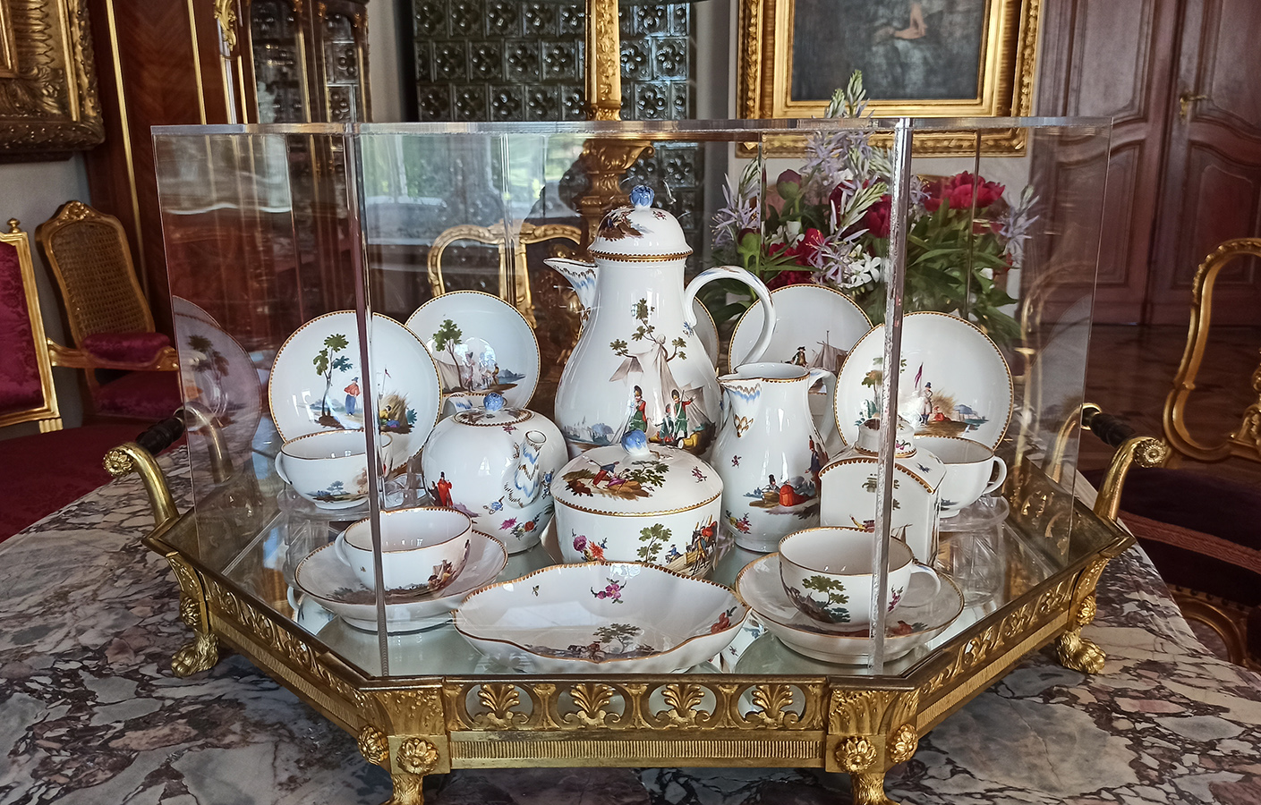 Serwis do herbaty i kawy ze scenami wojskowymi, Saksonia, 1763 – 1774; porcelana, farby naszkliwne, złocenia; zbiory Muzeum Zamoyskich w Kozłówce, nr inw. MPK/CS/1962-1988 Fotografia przedstawia serwis porcelanowy umieszczony na szklanej tacy z balustradką, nóżkami i uchwytami w złotym kolorze. Serwis w kolorze białym z kolorową dekoracją. W centrum wysoki dzbanek z pokrywką, przed nim niski pojemnik, z dwóch stron niższe dzbanki. Wkoło ustawione talerze i filiżanki. Na pierwszym planie miseczka. Serwis przykryty osłoną z przezroczystej pleksi. Taca stoi na marmurowym stole, z lewej strony widać fragment kanapy w kolorze bordowym z oparciami w kolorze złotym. Po prawej krzesło w kolorze bordowym z oparciem w kolorze złotym. w tle fragment wnętrza z piecem kaflowym, obrazami w ramach w kolorze złotym, z prawej strony w głębi drzwi.