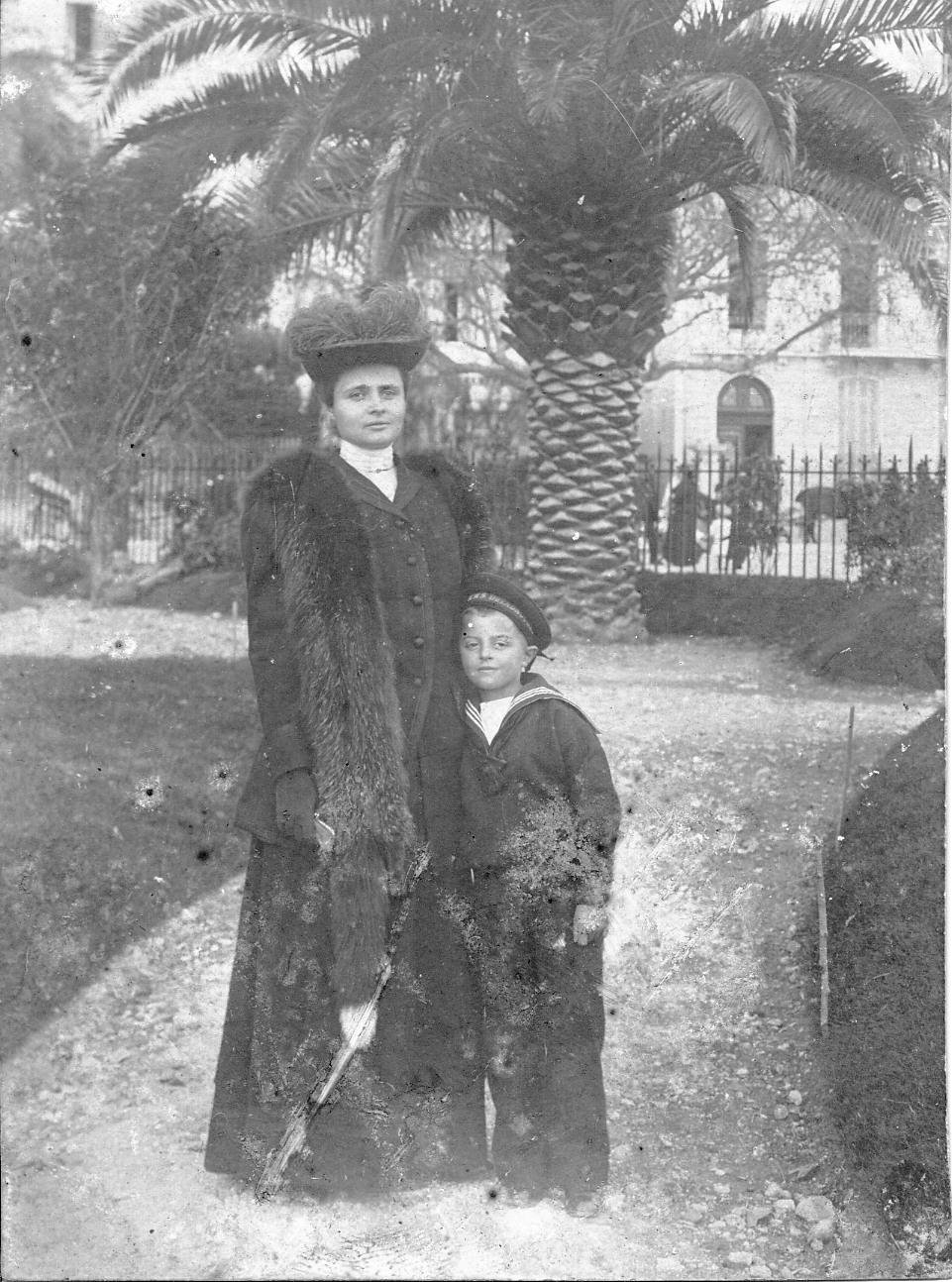 3. Maria z Potockich Adamowa Zamoyska z synem Michałem, Francja? Włochy?, ok. 1905 r. Fotografia archiwalna, czarno-biała, wykonana około 1905 roku, przedstawia Marię Potocką, późniejszą Adamową Zamoyską, z synem Michałem. Kobieta w czarnej, długiej spódnicy i żakiecie z narzuconą futrzaną etolą i bogato zdobionym kapeluszu. Przy niej, przytulony, stoi chłopiec, ubrany w ciemny, stylizowany na marynarski strój. W tle fragmenty budynków i okazała palma.