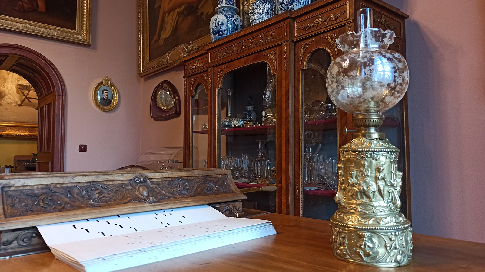 Fragment wnętrza Salonu Małego, stan obecny Fotografia kolorowa przedstawiająca fragment wnętrza Salonu Małego w pałacu kozłowieckim. Na blacie instrumentu ustawiona jest lampa naftowa o mosiężnym korpusie i szklanym, kwiatowym kloszu. Po lewej stronie widoczna perforowana papierowa taśma z zapisem muzycznym, w tle przeszklona witryna wypełniona przedmiotami, na ścianie oprawione w ramy obrazy i fragment drzwi.
