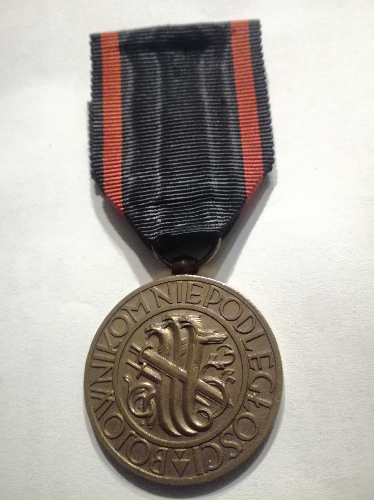 Medal Niepodległości (awers); kolekcja prywatna Fotografia Medalu Niepodległości, polskiego odznaczenia państwowego., Na wstędze orderowej w kolorze czarnym, z czewonym paskiem wzdłuż obu boków, zawieszony okrągły medal bity w brązie. W polu środkowym znajduje się wyobrażenie trzech hydr, przebitych trzema mieczami. W otoku jest napis: BOJOWNIKOM NIEPODLEGŁOŚCI.