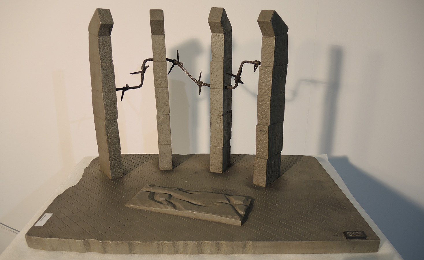 2. Miniatura pomnika ku czci pomordowanych w obozie KL Auschwitz III - Monowitz, lata 70. XX w., MPK/SW/2693 Fotografia miniatury pomnika ku czci pomordowanych w obozie KL Auschwitz III - Monowitz, powstałej latach 70. XX wieku. Na prostokątnej podstawie cztery słupy ogrodzenia obozowego, połączone drutem kolczastym. Przed nimi poziomo leżąca płyta z wyrzeźbionymi konturami postaci ludzkich. Na podstawie tabliczka z napisem: „Monowice – Oświęcim".