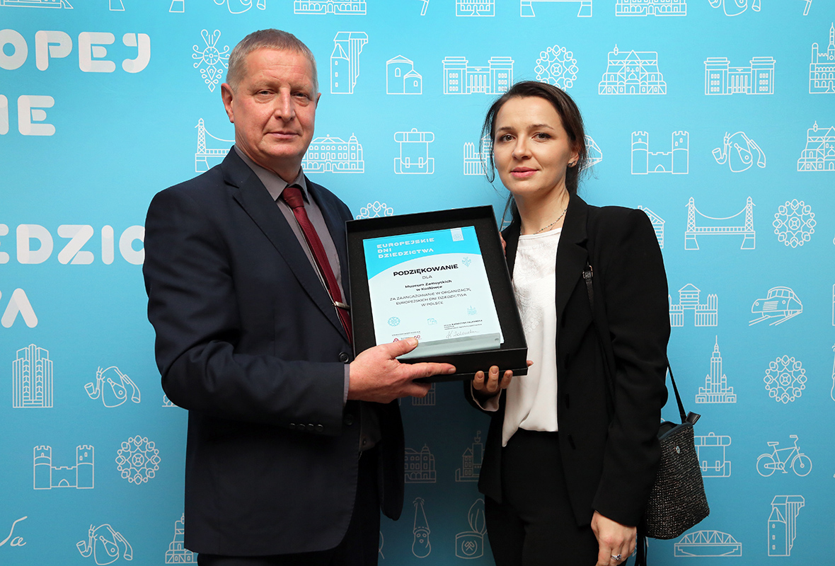 Pracownicy Muzeum Zamoyskich w Kozłówce odbierający wyróżnienie: Sławomir Grzechnik, Kierownik Działu Zbiorów Sztuki Współczesnej i Katarzyna Kot, kustosz; fot. Paweł Kobek (NID) Fotografia kolorowa przedstawia pracowników Muzeum Zamoyskich w Kozłówce odbierających wyróżnienie Narodowego Instytutu Dziedzictwa: Sławomira Grzechnika, Kierownika Działu Zbiorów Sztuki Współczesnej i Katarzynę Kot, kustosza; na tle błękitnej ścianki z logotypami EDD i fragmentem napisu mężczyzna i kobieta, ubrani w ciemne garnitury trzymają pamiątkową plakietę; fot. Paweł Kobek (NID)