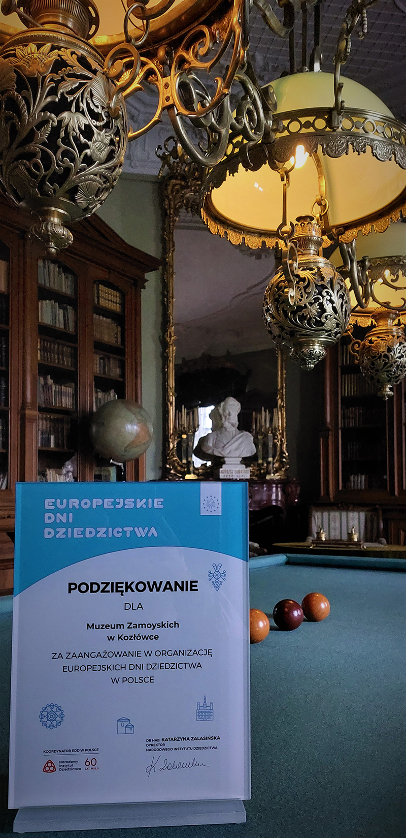 Plakieta pamiątkowa dla wyróżniających się instytucji. Fotografia kolorowa przedstawia plakietę pamiątkową dla instytucji zaangażowanych w organizację Europejskich Dni Dziedzictwa w Polsce, przyznaną Muzeum Zamoyskich w Kozłówce przez Narodowy Instytut Dziedzictwa; Na pierwszym planie plakieta pamiątkowa z logotypami i napisem „Europejskie Dni Dziedzictwa Podziękowanie dla Muzeum Zamoyskich w Kozłówce za zaangażowanie w organizację Europejskich Dni Dziedzictwa w Polsce” z odręcznym podpisem dr hab. Katarzyny Zalasińskiej, dyrektora Narodowego Instytutu Dziedzictwa.