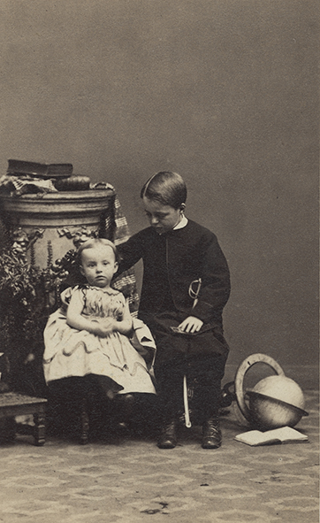 Władysław i Maria Zamoyscy. Fotografia przedstawia dwoje dzieci. Dziewczyna – Maria siedzi na niskim krzesełku. Ma jasne, gładko uczesane włosy z przedziałkiem na środku. Jest ubrana w jasną sukienkę. Na kolanach trzyma splecione rączki. Obok na siedzi chłopiec – Władysław. Ma ciemne włosy, uczesane gładko z przedziałkiem z prawej strony. Ubrany jest w ciemny żakiet i spodnie, przy szyi widać biały kołnierzyk. Prawą rękę ma opartą o krzesło na którym siedzi jego siostra; lewą opartą na kolanach. Patrzy w kierunku dziewczynki. Z tyłu za nimi stoi komoda, na której leżą książki. Obok, na podłodze leży globus.