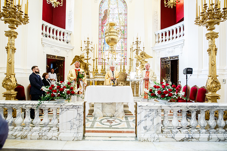 Obchody jubileuszu 80-lecia Muzeum Zamoyskich w Kozłówce. Zdjęcie przedstawia bogato zdobione wnętrze barokowego kaplicy w Kozłówce. Na środku znajduje się ołtarz z białym obrusem, przy którym stoi biskup w złotej szacie liturgicznej oraz dwóch innych księży w białych i czerwonych strojach. W tle widoczne są złote kandelabry, anioły, oraz kolorowe witraże. Po lewej stronie stoją dodatkowe osoby w eleganckich ubraniach.