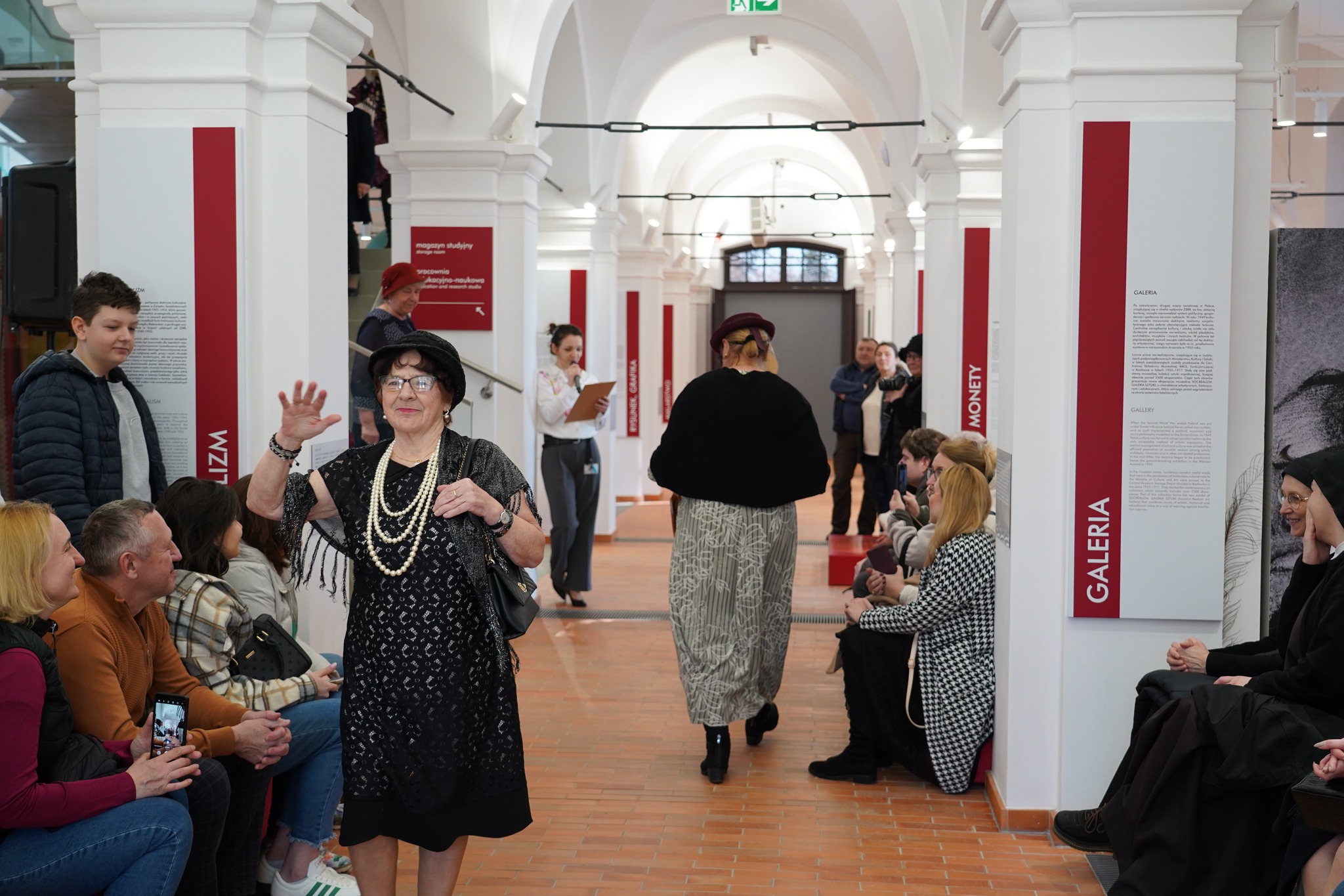Muzeum na obcasach - Dzień Kobiet w Muzeum Zamoyskich Na zdjęciu w eleganckim, jasnym wnętrzu z białymi, sklepionymi sufitami i czerwono-białymi tablicami informacyjnymi na ścianach trwa wydarzenie przypominające pokaz mody.
Na pierwszym planie starsza kobieta w czarnej, koronkowej sukni z frędzlami, ozdobiona długimi perłowymi naszyjnikami i bransoletami, kroczy z uśmiechem. Ma na sobie czarny kapelusz, okulary i trzyma torebkę na ramieniu. Jej prawa ręka jest uniesiona w geście powitania lub prezentacji.
Za nią inna kobieta w stylowym stroju z długą spódnicą i czarnym futrzanym okryciem odchodzi w głąb wybiegu. Na dalszym planie widać prowadzącą wydarzenie osobę w białej koszuli i szarych spodniach, trzymającą notatki.
Publiczność, ubrana w codzienne stroje, siedzi po obu stronach wybiegu. Niektórzy uśmiechają się, inni patrzą z zainteresowaniem, a jedna osoba nagrywa wydarzenie na telefonie. Wśród widzów widać także zakonnicę w czarnym habicie, która rozmawia z inną kobietą.
Atmosfera zdjęcia wskazuje na kulturalne wydarzenie, być może pokaz mody retro lub prezentację historycznych strojów, odbywającą się w muzealnej lub wystawienniczej przestrzeni.
