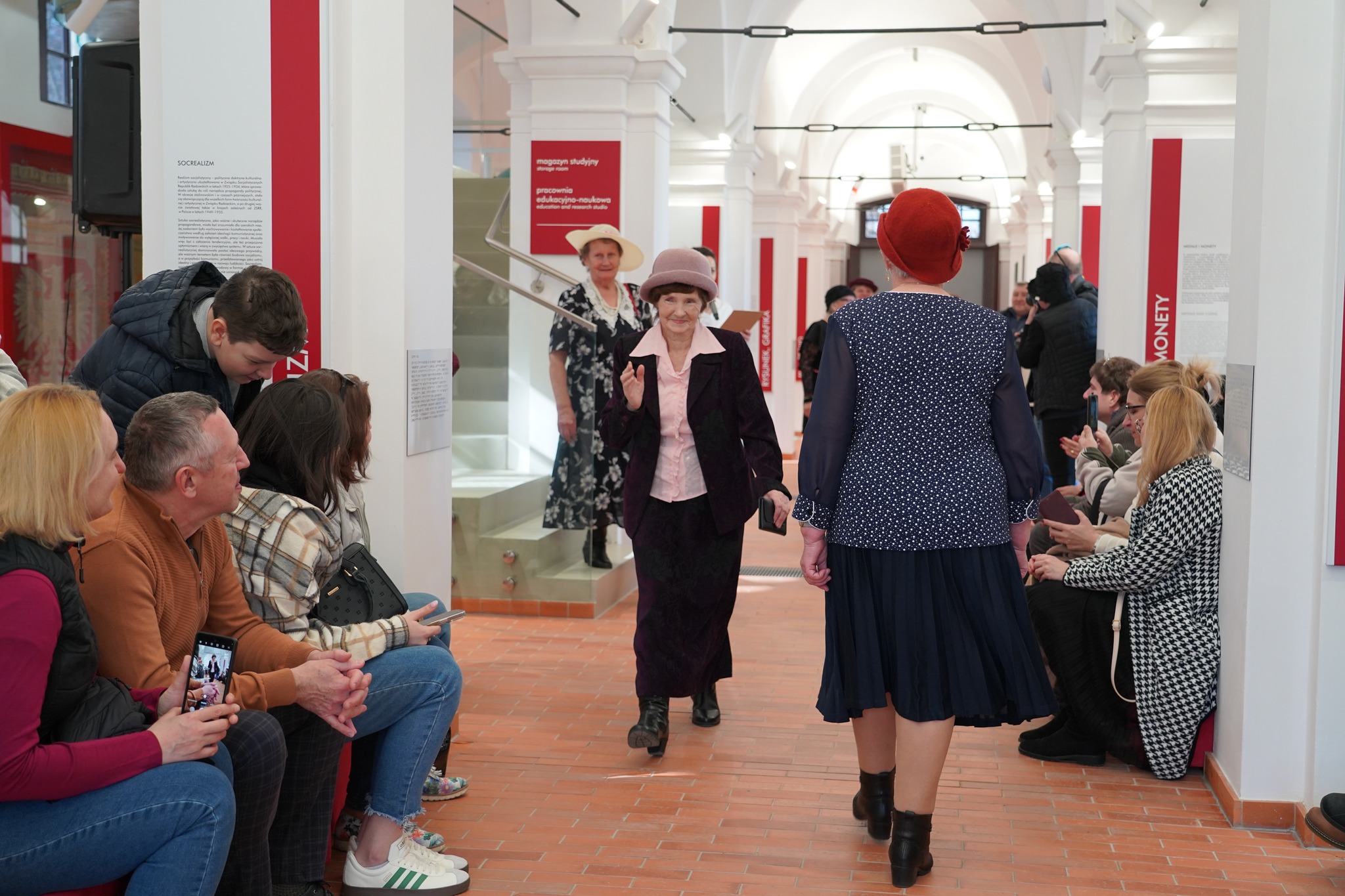 Muzeum na obcasach - Dzień Kobiet w Muzeum Zamoyskich Na zdjęciu w przestrzeni wystawienniczej lub muzealnej odbywa się pokaz mody retro. Wnętrze charakteryzuje się białymi, sklepionymi sufitami i czerwono-białymi tablicami informacyjnymi na ścianach.
W centrum kadru starsza kobieta w eleganckim, bordowym garniturze i różowej koszuli zbliża się w stronę publiczności. Na głowie ma pasujący kapelusz, a w ręce trzyma małą kopertówkę. Uśmiecha się delikatnie i unosi dłoń w geście powitania.
Na pierwszym planie widoczna jest inna kobieta, idąca w przeciwnym kierunku, ubrana w granatową sukienkę w białe groszki, z długim rękawem i plisowanym dołem. Na głowie ma czerwony beret, a na nogach czarne botki na obcasie.
W tle, na schodach, stoi kolejna uczestniczka pokazu – ubrana w długą, czarną sukienkę z białym wzorem i jasny kapelusz. Dalej, przy wejściu do sali, zgromadzona jest grupa widzów i organizatorów.
Po obu stronach wybiegu siedzi publiczność, składająca się z osób w różnym wieku. Niektórzy uśmiechają się, inni robią zdjęcia telefonami. Po prawej stronie widać kobietę w czarno-białym płaszczu w pepitkę oraz osoby w okularach i ciepłych ubraniach.
Zdjęcie ukazuje przyjazną, pełną zainteresowania atmosferę, sugerując, że jest to pokaz mody historycznej lub stylizowany na dawne epoki, odbywający się w przestrzeni kulturalnej.