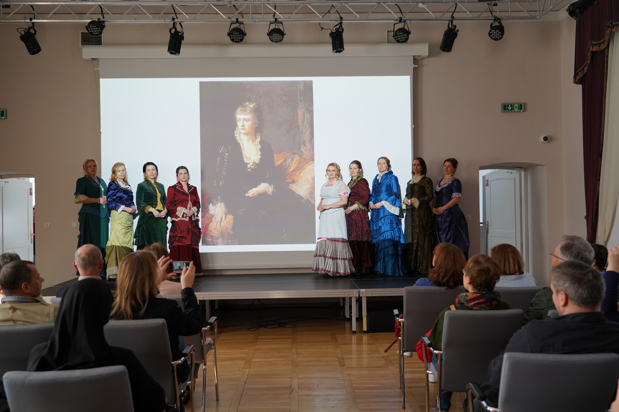 Muzeum na obcasach - Dzień Kobiet w Muzeum Zamoyskich Fotografia przedstawia pokaz mody XIX-wiecznej. Dziewięć modelek, młodych kobiet, stoi na podwyższonej scenie w dużym, przestronnym pomieszczeniu, cztery po lewej stronie oraz pięć po prawej stronie. W centralnej części znajduje się duży ekran z wyświetlonym wizerunkiem obrazu przedstawiającego kobietę ubraną według mody XIX-wiecznej. Na pierwszym planie widać od tyłu publiczność wydarzenia, widać siedzących na szarych krzesłach grupę ludzi po prawej i lewej stronie.
Modelki, od lewej: kobieta w zielonej ciemnozielonej długiej sukni z rękawami ¾. Na szyi zawieszone ma białe perły sięgające do pasa. Włosy ma krótko upięte i udekorowane białą delikatną opaską. Druga od lewej kobieta ubrana jest w dwuczęściowy kostium, żółtą długą spódnicę z dekoracyjnym dołem oraz granatową bluzkę, obie części stroju są aksamitne. Kolejna kobieta ubrana jest w dwuczęściowy ciemnozielony strój, długą spódnicę oraz krótką marynarkę, spod której wystaje żółto-złota bluzka. Kolejna kobieta, ubrana w bordowy kostium, długą bordową spódnicę (upiętą na wysokości kolan) oraz bordową marynarkę z eleganckim kołnierzem wykończonym białą koronką oraz podobnie wykończonymi rękawami.