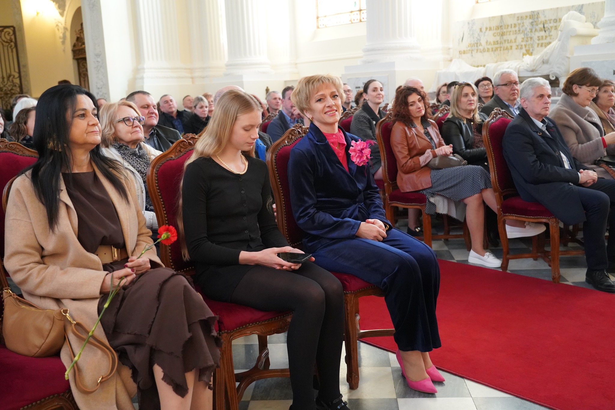 Muzeum na obcasach - Dzień Kobiet w Muzeum Zamoyskich Fotografia przedstawia wnętrze kaplicy pałacowej podczas koncertu Kameralnej Orkiestry Akordeonowej „Arti Sentemo” i wskazuje na publiczność wydarzenia. Wnętrze kaplicy jest jasne, rozświetlone, przestronne, elementy architektoniczne wykonane są z białego marmuru. Wszystkie miejsca w kaplicy pałacowej są zajęte. Na pierwszym planie widnieją trzy kobiety, od lewej kobieta w średnim wieku z ciemnymi włosami, jasną karnacją i ciemną oprawą oczu, ubrana elegancko. Ma na sobie brązową sukienkę przepasaną w pasie beżowym paskiem oraz jasno-beżowy płaszcz. Obok niej na krześle leży mała ciemno-beżowa torebka. Obok niej, po prawej stronie siedzi młoda dziewczyna o naturalnej , delikatnej urodzie, jest szatynką z jasną karnacją i szatynowi włosami. Ubrana jest cała na czarno. Ma rajstopy, krótką spódniczkę oraz sweterek. Po prawej stronie siedzi kobieta w średnim wieku, o krótkich jasnych włosach, delikatnej urodzie i filigranowej figurze. Ma na sobie aksamitny granatowy komplet, spodnie w kant i marynarkę. Do klapy marynarki ma przypięty duży różowy kwiat w formie broszki.